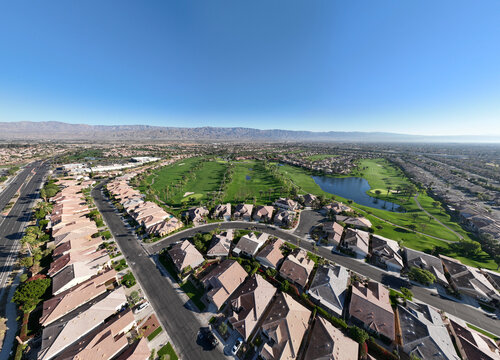 Aerial view of a residential neighborhood featuring a green golf course with a pond, tiled roof houses, and distant mountains under a clear blue sky in Indio, California, United States.