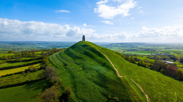 Aerial view of Glastonbury Tor and St Michael's Tower rising above the lush green Somerset countryside under a bright blue sky in Glastonbury, England, United Kingdom.