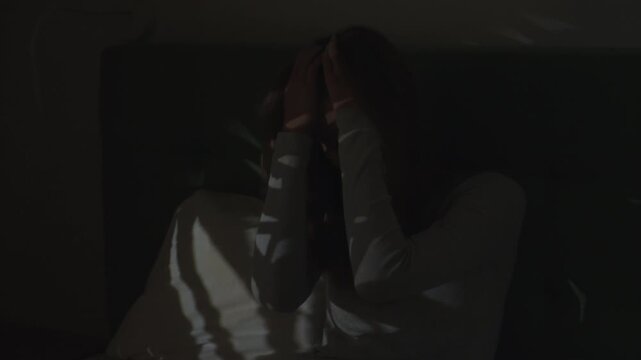 Woman in a dark bedroom. Lying in bed despairing with face in hands and looking outside through the window. Suffering from depression