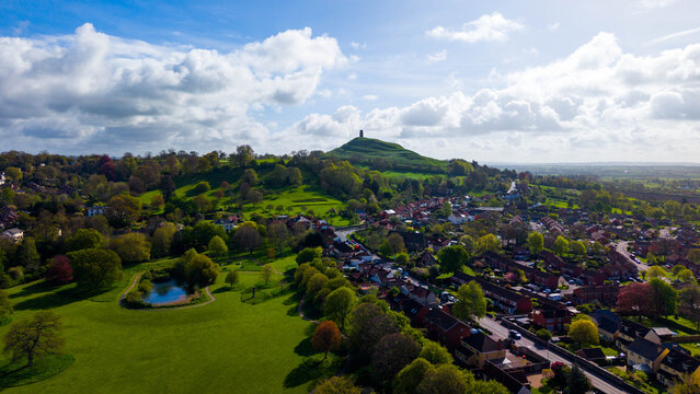 Aerial view of Glastonbury Tor rising above the town of Glastonbury with lush green fields, trees, and residential houses under a bright blue sky in Glastonbury, England, United Kingdom.