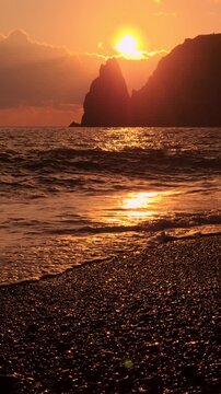 Golden sunset ocean waves wash over a rocky pebble beach near dramatic cliffs. Static eye-level shot.