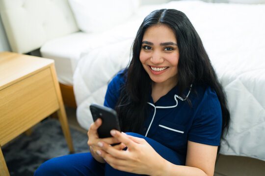 Happy young woman sitting on bedroom floor wearing pajamas, smiling and relaxing while using a smartphone