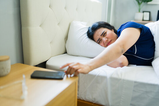 Latin woman reaching for smartphone alarm, feeling sleepy and tired in bed, waking up in the morning