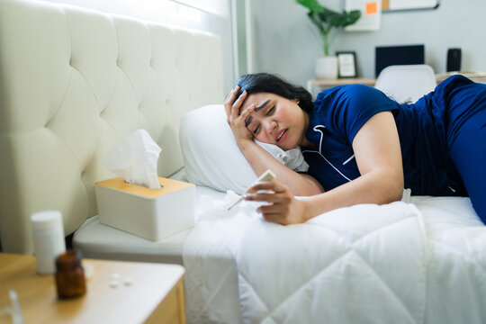 Woman suffering from fever and headache, lying sick in bed at home, checking thermometer for high temperature