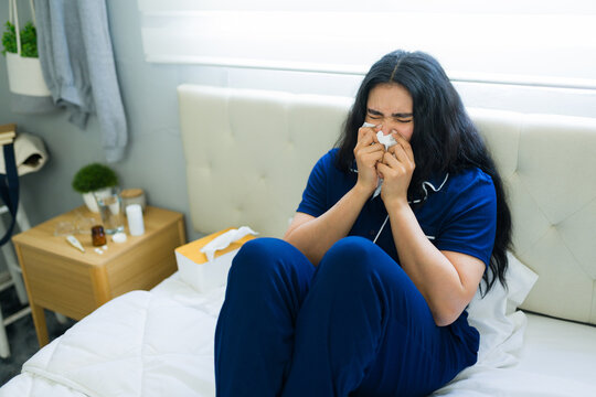 Young woman blowing nose in bed feeling unwell with cold or flu, dealing with allergies or sickness at home