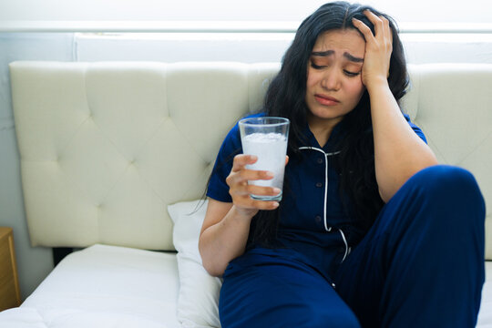 Hungover woman suffering from headache in bed, holding glass of water for relief, experiencing pain and illness at home