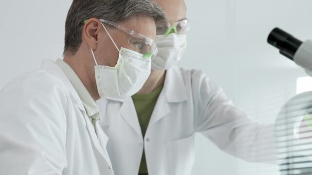 Two diverse scientists. Wearing lab coats. Safety goggles. And masks. Performing detailed research using a microscope in a clean laboratory setting