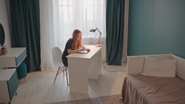 Young woman with long red hair sits at white desk. Room features teal walls and dark green curtains natural light filters. Bed with beige bedding is visible on one side of scene