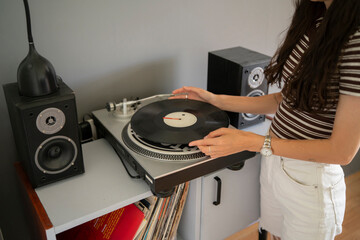 A person holds a vinyl record above a turntable in a room with audio speakers and a shelf of...