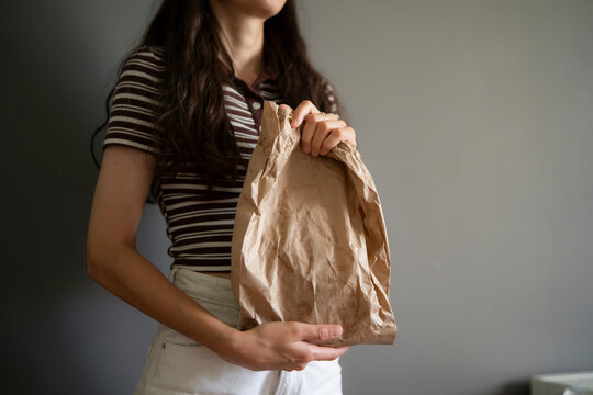 A person stands indoors holding a brown paper bag while wearing a striped shirt and light pants