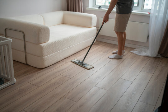 A person mops the floor in a simple living room with a couch and a window during the day. The space is tidy