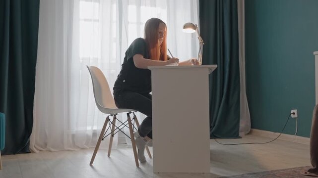 Young woman with long hair seated at white desk writing. Woman interacts with pen or small object while remaining focused. White chair has wooden legs gold table lamp is lit
