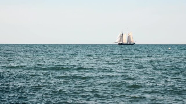 Traditional sailing ship crosses calm Baltic Sea under pale sky, white sails glowing in soft sunlight. Minimal maritime scene highlights horizon, open water, navigation, travel and peaceful motion