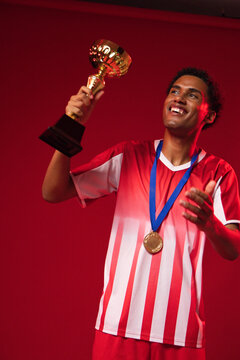 Adult male athlete wearing red-and-white striped jersey and gold medal raising trophy in studio