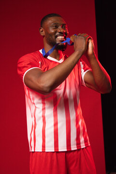 African man holding gold medal on blue ribbon near mouth in red studio wearing striped jersey