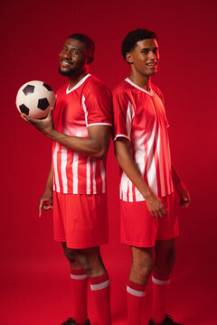 African American male players standing back-to-back in studio holding soccer ball, wearing red kits