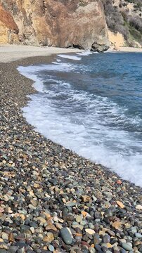 Ocean waves crash against colorful pebble stones on a remote beach shoreline near tall cliffs. Low angle static shot.