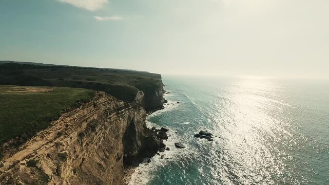 Aerial view captures cliff edge meeting ocean. Green grass blankets cliffside. Sunlight glints off calm sea waters. Sky shows soft clouds above. Wind rustles through grass gently
