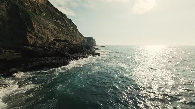 Coastal cliffs meet ocean waves crashing against rocky shores. Sunlight glints on turquoise waters under partly cloudy skies. Waves surge with white foam along rugged, mossy cliffsides