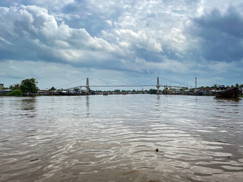 Wide suspension bridge spans murky river under cloudy sky. Boat's wake ripples across brown water beneath. Structural towers anchor bridge with taut cables.