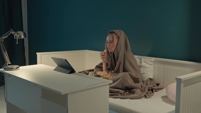 Young woman sits on bed covered with brown blanket while eating chips. Chips in clear container silver lamp illuminates white desk where. Teal wall background with minimal decor