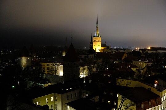 high-angle view of Tallinn Old Town at night, featuring medieval city wall defense towers and the illuminated St. Olaf's Church spire