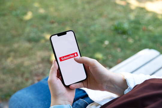 Subscribe icon. Woman sitting on a park bench with a smartphone, tapping the social media subscribe button on the screen. Internet, newsletter, e-commerce, mobile app, wireless technology.