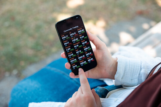 Woman sitting on a park bench with a smartphone, tapping  on the screen with commodity trading information. Stock market and exchange, internet, mobile app, wireless technology.