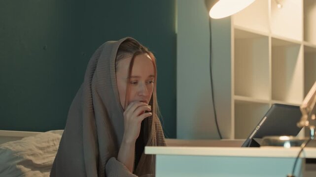 Young woman sits at desk covered by gray blanket over her head and shoulders. Woman interacts with electronic device. Background features teal walls white shelving units