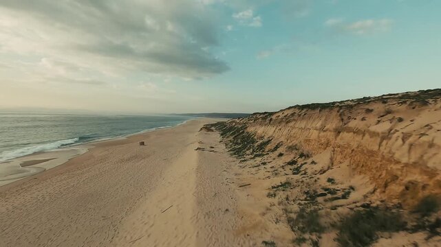 Aerial view of serene beach with sandy dunes and ocean waves. Path winds through dunes toward water. Calm sky with scattered clouds above. Ideal for travel, nature, or vacation marketing