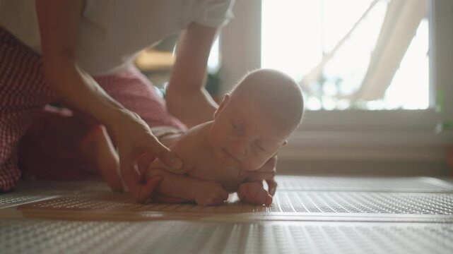 Adorable newborn baby boy with bright blue eyes doing tummy time on a mat, with his mother's gentle hands supporting him as he learns to lift his head and strengthen his neck muscles