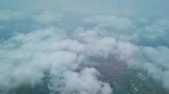 The aerial view from an airplane shows a thick layer of white clouds above a patchwork of agricultural fields. Faint sections of the landscape remain visible through openings between the clouds.