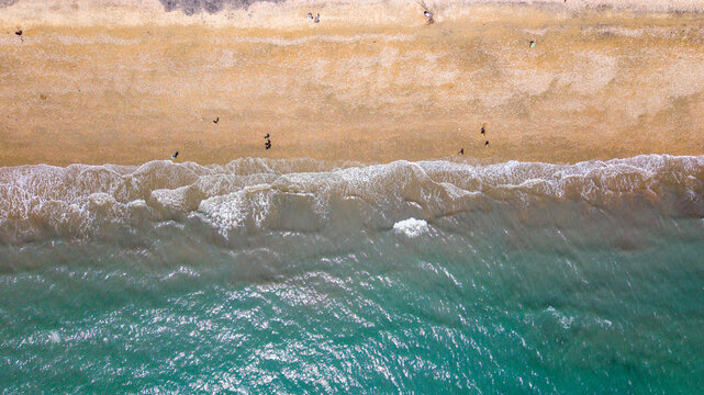 Aerial view of West Bay beach along the Jurassic Coast with gentle waves washing onto the golden sand and people walking along the shore in Bridport, Dorset, United Kingdom.