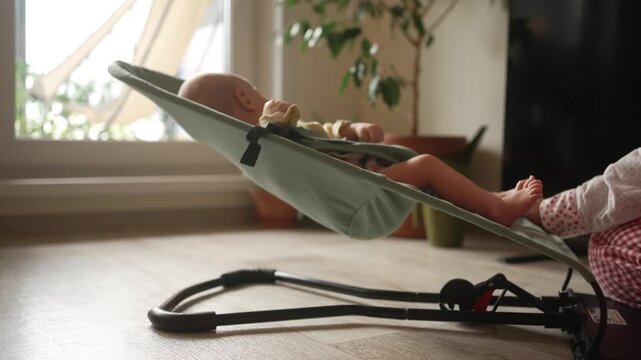 Sweet older sister gently rocks her newborn sibling in a bouncer on the living room floor, a tender moment of sibling affection and quiet domestic life as the infant gently sways
