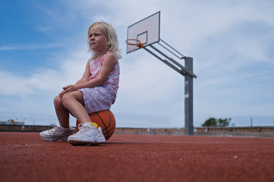 Cute Blonde Toddler Dribbling a Basketball Near a Hoop on an Outdoor Court