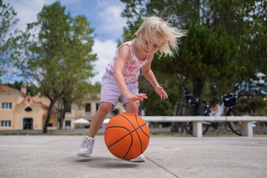 Cute Blonde Toddler Dribbling a Basketball Near a Hoop on an Outdoor Court