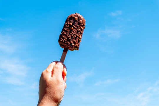 Hand holding crisp chocolate ice cream bar with nuts on wooden stick against clear blue sky in summer. Delicious cold sweet dessert snack outdoors on bright sunny day. Ideal for refreshment design