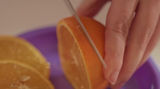 Orange is being sliced by steady hand motion. Fingers grip orange steady while blade cuts through rind. Purple plate or board serves as cutting area for fruit preparation