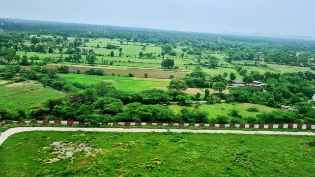 The aerial view shows a lush rural landscape with green agricultural fields, scattered trees and homes, and a winding paved path. Hazy hills form a distant horizon beyond the greenery.
