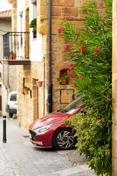 Modern red hatchback parked on a narrow European village street with vibrant pink oleander flowers in the foreground