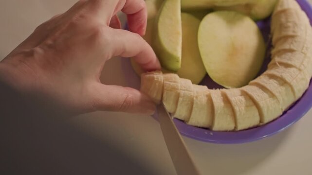 Close up of hand slicing peeled banana into small pieces. Hand arranges sliced bananas in circular pattern while knife remains. Neutral background emphasizes food preparation area