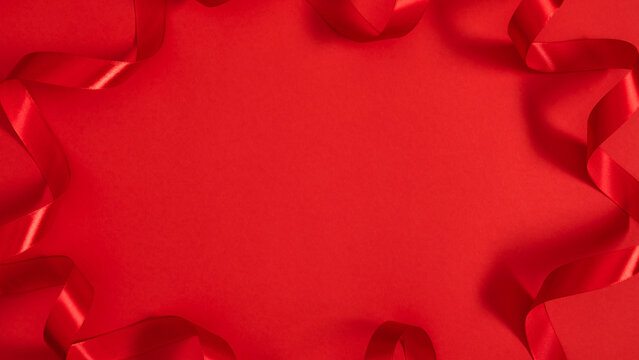 Vibrant red background with wide central copy space. Elegant border frame of wavy satin ribbon curls on red paper backdrop. Horizontal flat lay composition for Valentine Day greeting or Christmas sale