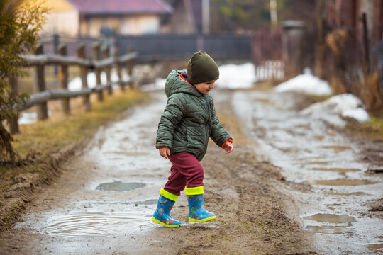 Little boy in protective rubber boots and rain clothes jumping in mud puddle. Happy child having fun while playing in puddle after rain.