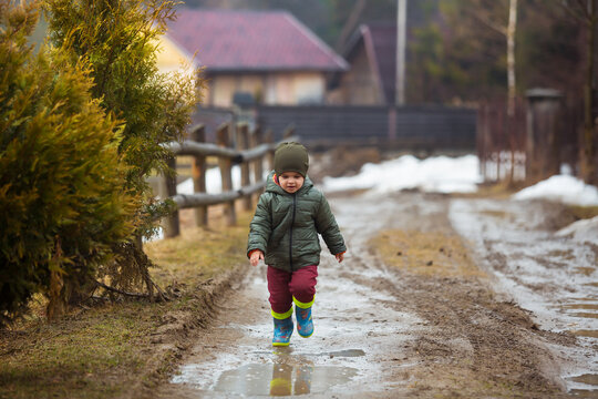 Little boy in protective rubber boots and rain clothes jumping in mud puddle. Happy child having fun while playing in puddle after rain.
