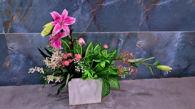 The colorful artificial floral arrangement features pink lilies, small white and pink blossoms, and patterned green foliage. A white square vase stands against a dark blue marble wall.