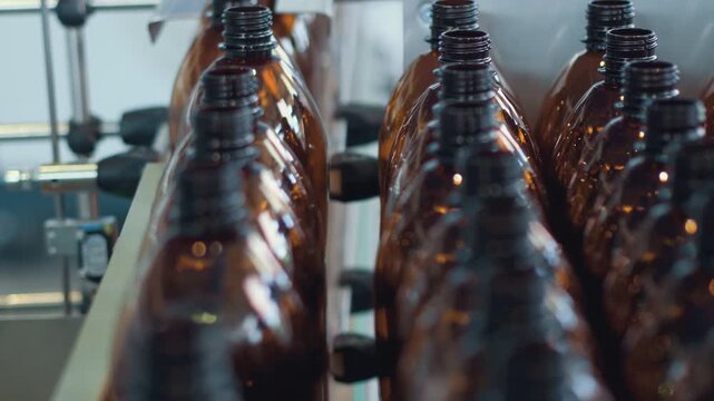 Rows of amber glass bottles on conveyor guides inside bright bottling facility, showing industrial packaging line, threaded necks, reflections, symmetry, depth, and orderly repetition