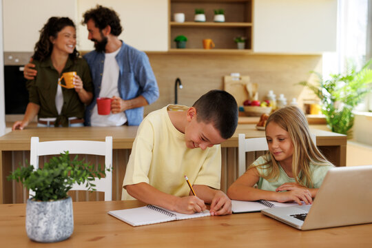 Children focusing on schoolwork and online learning at a kitchen table, with their parents standing in the background, enjoying morning coffee and a happy family moment