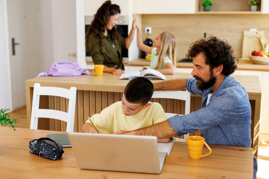 Father helping his son with schoolwork at a kitchen table while the mother and daughter celebrate an achievement in the background, showing family education and support
