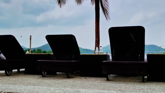 Two dark wicker lounge chairs frame the palm-lined lakeside view beside a small table and ornamental grass. Verdant hills rise beyond the water under a pale cloudy sky in Udaipur.