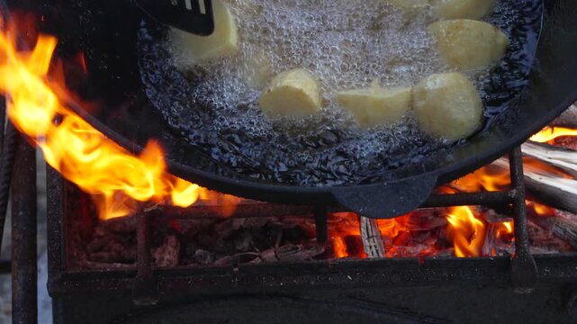 Sizzling potato chunks frying inside a cast iron wok over an open wood fire. High angle shot.